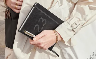 Person holding a black 2023 diary in a beige trench coat with a gold bracelet and rings.