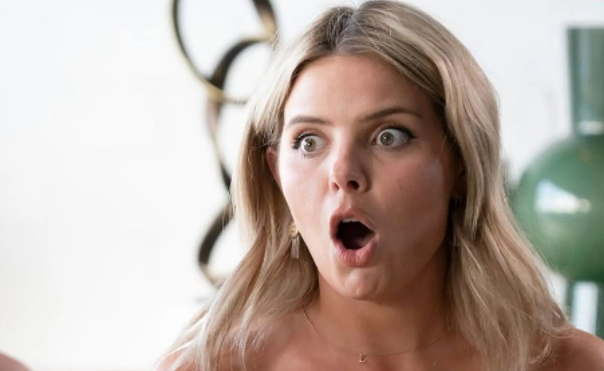 A woman with blonde hair looks surprised, mouth open and eyes wide, in a bright, modern setting.