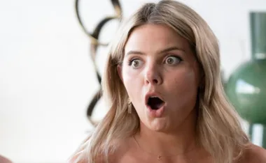 A woman with blonde hair looks surprised, mouth open and eyes wide, in a bright, modern setting.