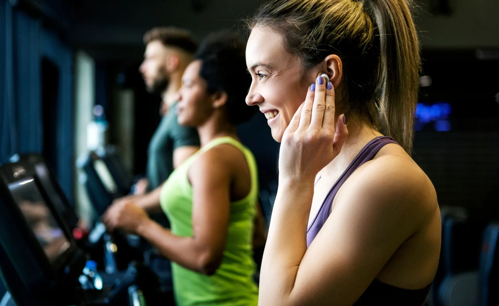 Woman using wireless earbuds on a treadmill, smiling at the gym with others exercising.