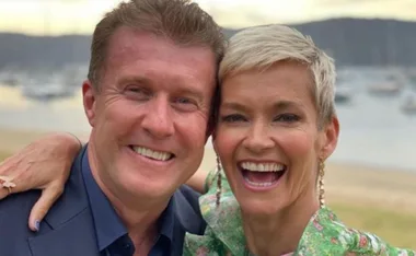 A couple smiling broadly, with a scenic waterfront background and boats in the distance.