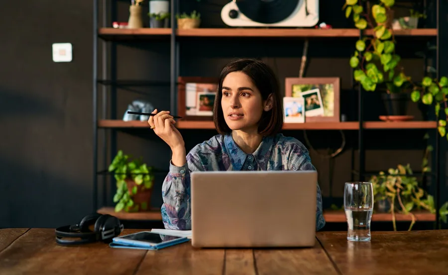 Person working at a desk with a laptop, pen in hand, surrounded by plants and shelves, focused and thoughtful.