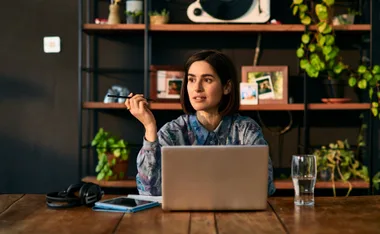 Person working at a desk with a laptop, pen in hand, surrounded by plants and shelves, focused and thoughtful.