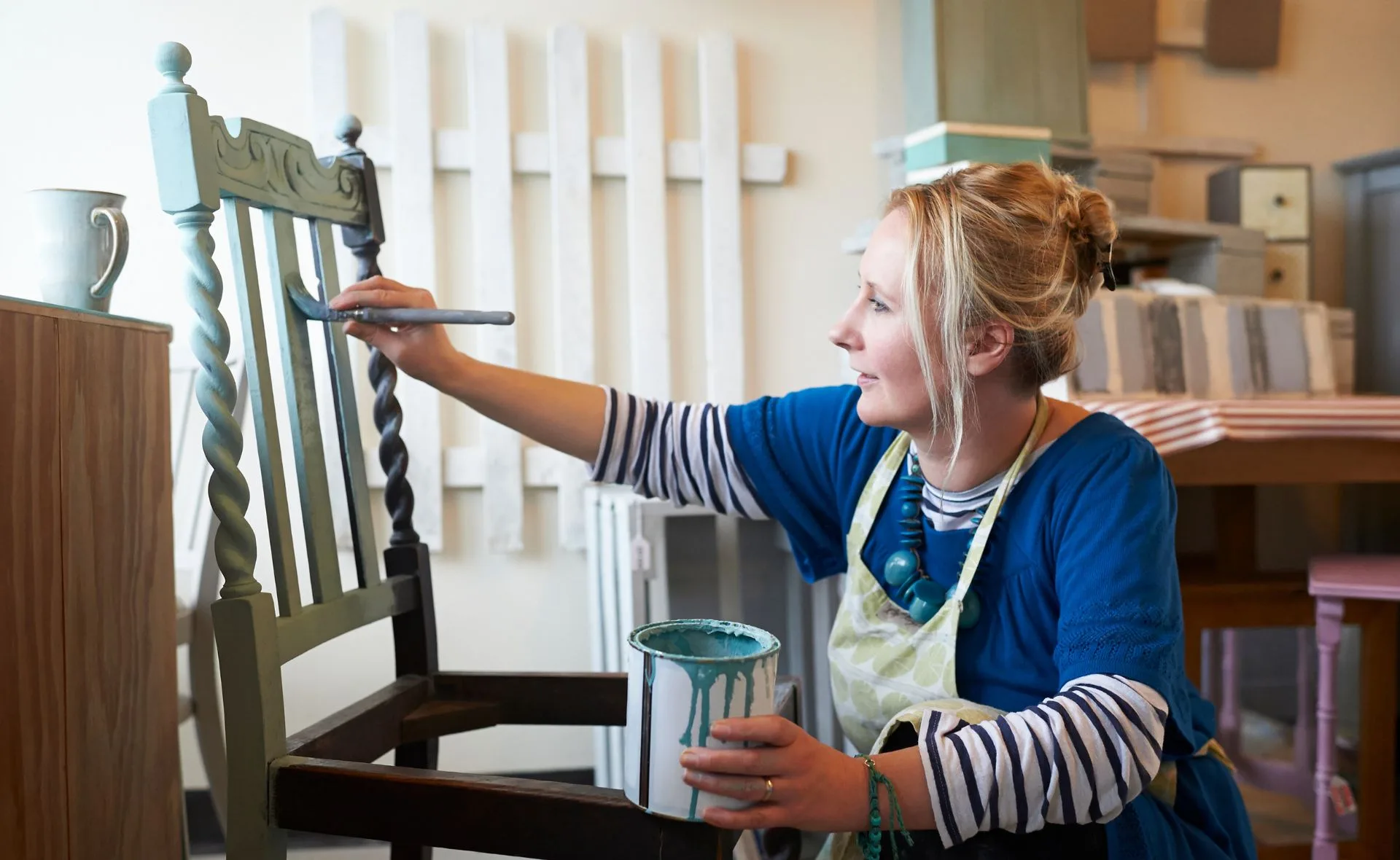 A woman in a workshop painting an old chair with blue paint.