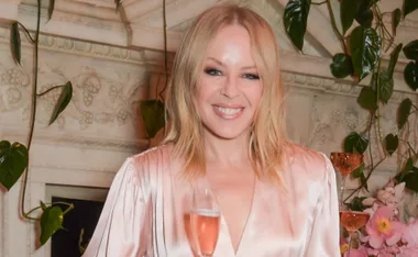 A woman in a silky pink dress smiles, holding a glass of pink champagne, with floral decor in the background.