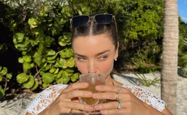 Woman with sunglasses on head drinking from a glass outdoors, surrounded by greenery.