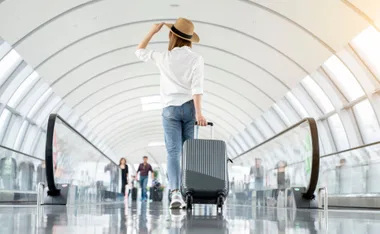 Traveler in a white shirt and hat pulling a gray suitcase through a modern, arched airport terminal.