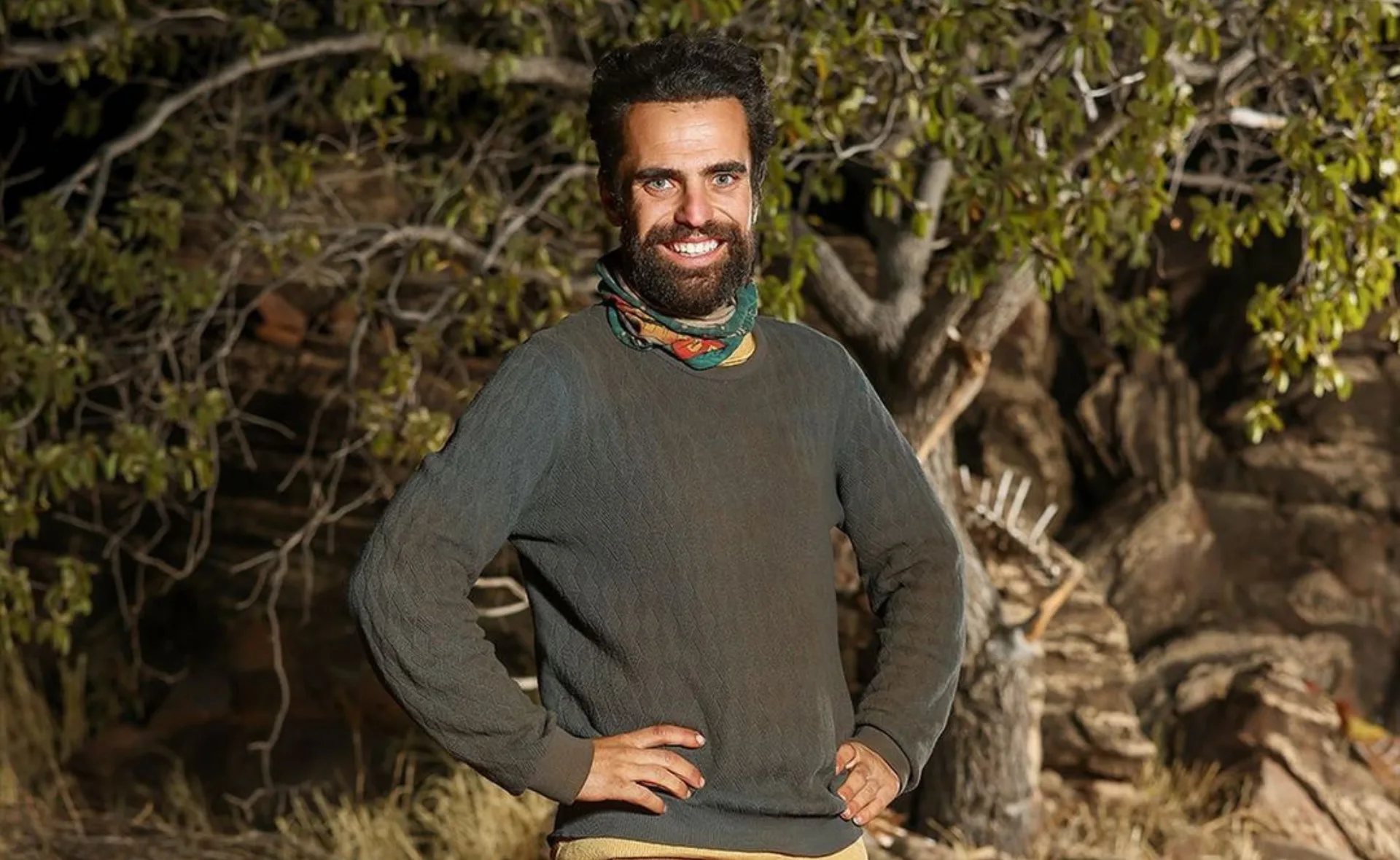 Man in a dark sweater smiling with hands on hips, standing outdoors in front of a tree.