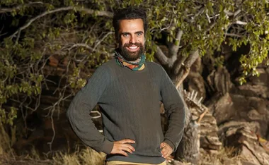 Man in a dark sweater smiling with hands on hips, standing outdoors in front of a tree.