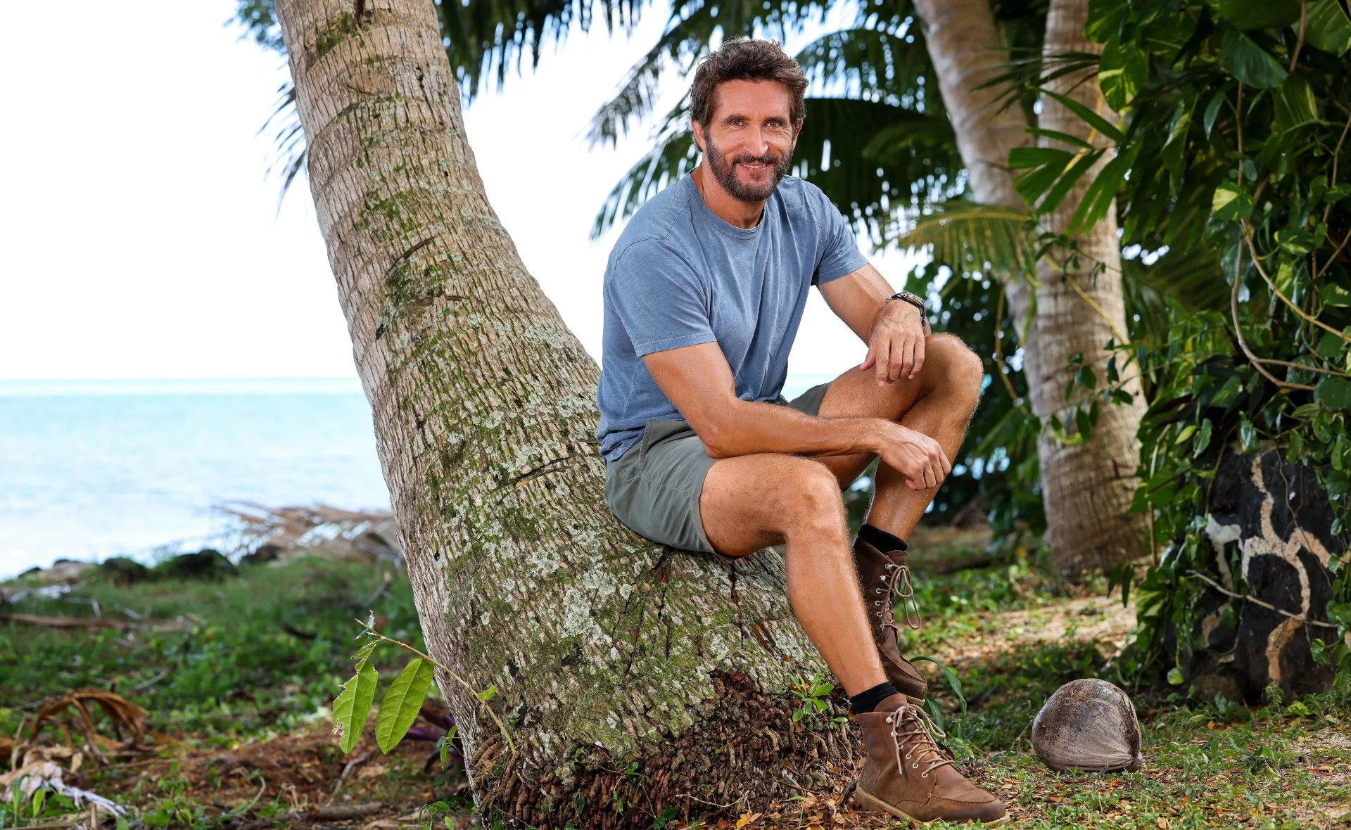 Man in a blue shirt and shorts sitting on a leaning tree by the beach, surrounded by greenery.