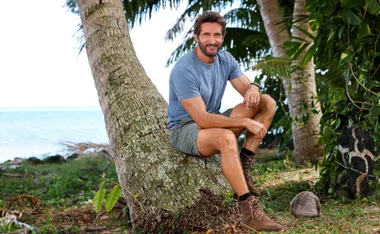 Man in a blue shirt and shorts sitting on a leaning tree by the beach, surrounded by greenery.