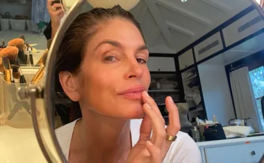 A person applying lip balm while looking into a mirror, with makeup brushes in the background.