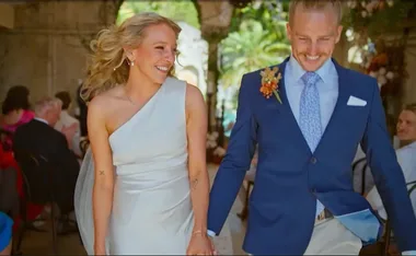 Couple smiling and walking hand in hand at a wedding ceremony, with guests seated in the background.