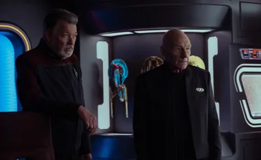 Two men in a futuristic room with glowing blue and gold artifacts behind them.