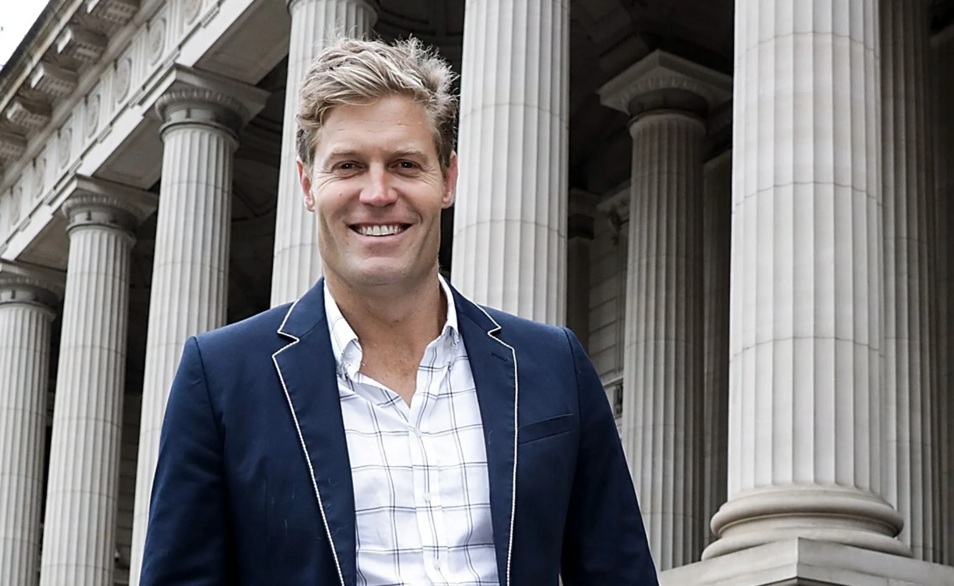 Man in a suit smiling in front of a building with large columns.