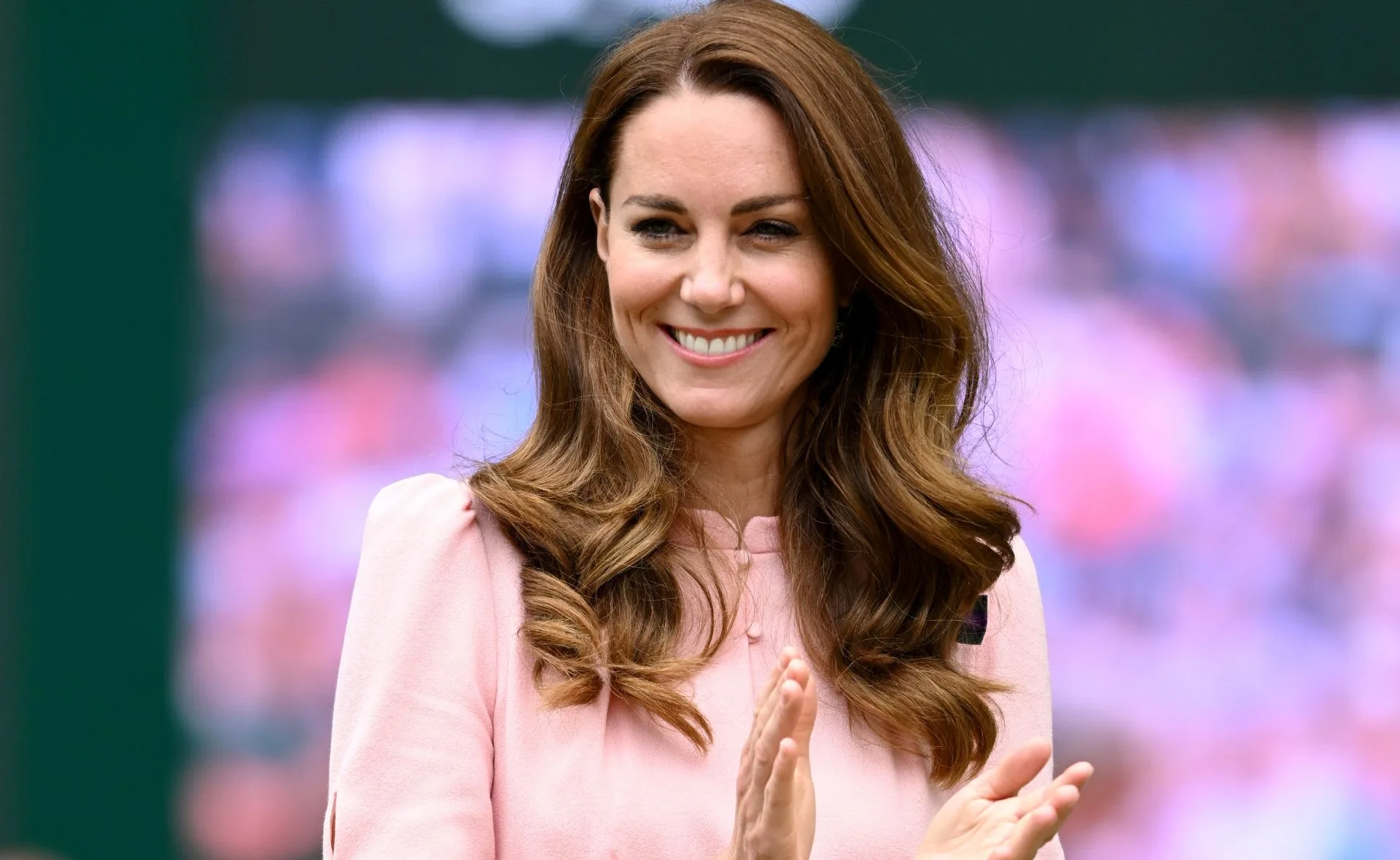 Clapping woman in a pink dress, smiling, with a blurred background.