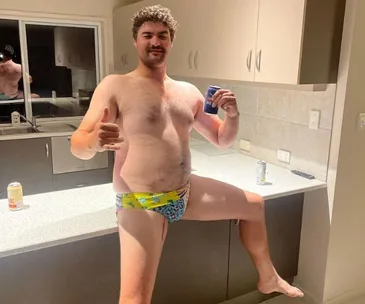 Man in colorful swim briefs posing in a kitchen with a drink, giving a thumbs-up sign.