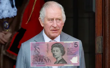 King Charles standing, holding an Australian five-dollar note featuring Queen Elizabeth II's portrait.