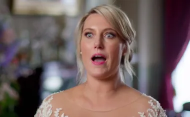 A woman in a white lace dress looks shocked with open mouth indoors.