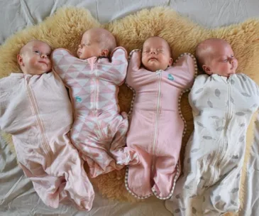 Four newborns in soft sleepers lie side by side on a fluffy rug, each wrapped in a differently patterned swaddle.