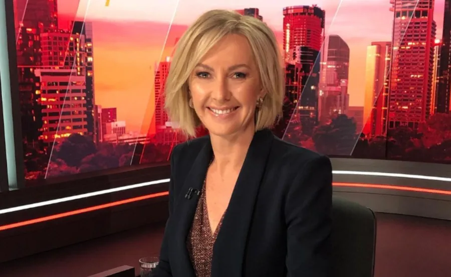 Smiling person in a blazer on a news set with a cityscape backdrop.