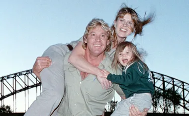 A smiling family poses together outdoors, with a bridge in the background at dusk.