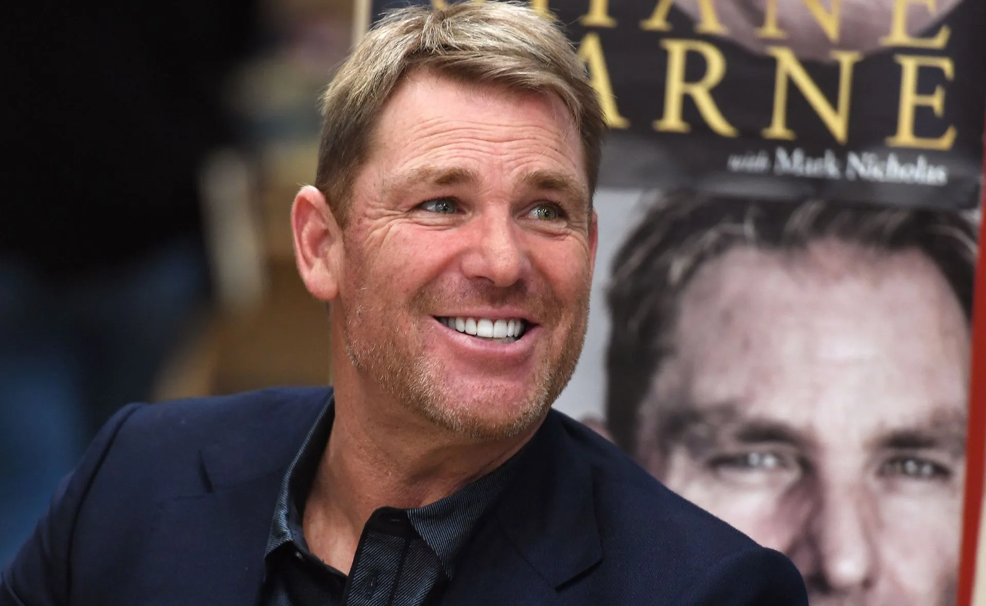 Man smiling at a book signing event, book cover featuring his face in the background.