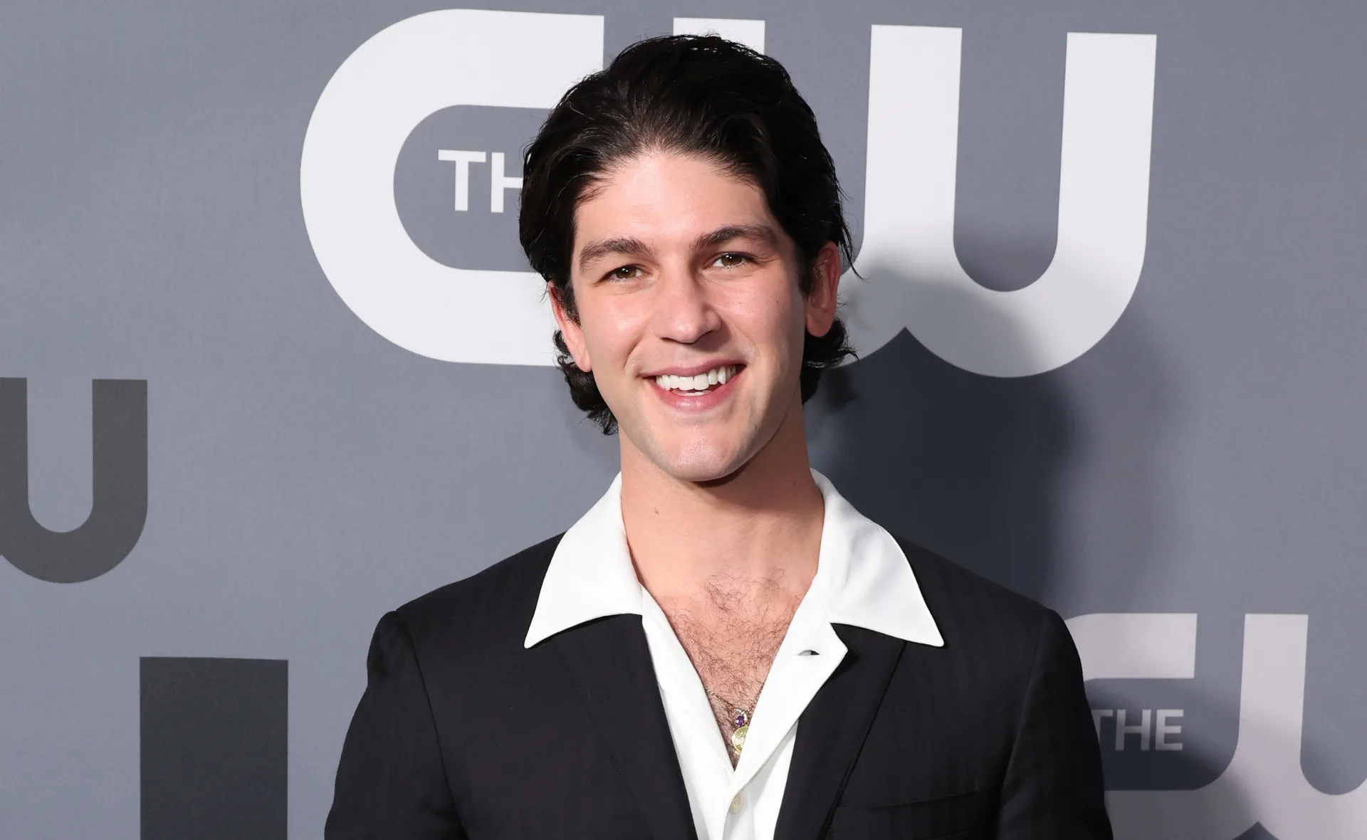 Smiling person in a black suit with a white shirt in front of a CW backdrop.