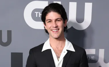 Smiling person in a black suit with a white shirt in front of a CW backdrop.