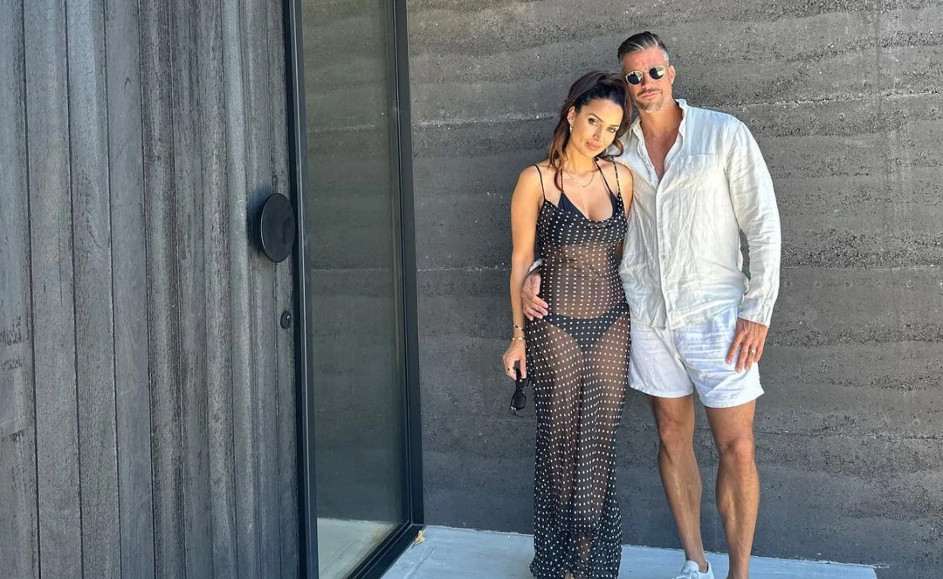 Couple posing in summer attire outdoors by a modern building, one in a sheer dress and the other in a white outfit.