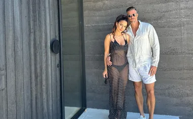 Couple posing in summer attire outdoors by a modern building, one in a sheer dress and the other in a white outfit.