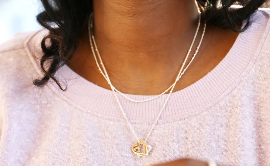 A woman wearing layered gold chains and a heart-shaped pendant necklace over a light pink sweater.
