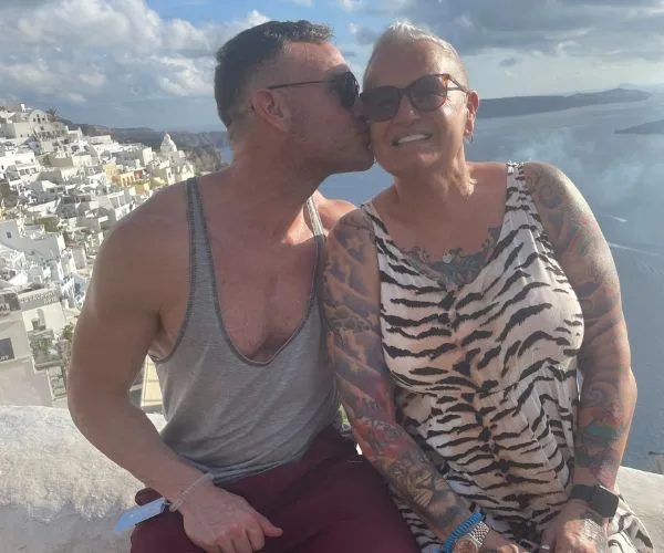 Man kissing woman on the cheek, both smiling, with a coastal town and ocean in the background.