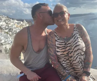 Man kissing woman on the cheek, both smiling, with a coastal town and ocean in the background.