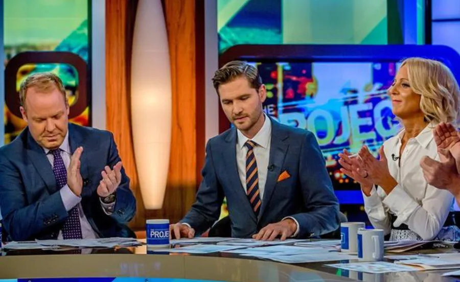 A TV studio set with three hosts in suits, clapping and discussing papers.