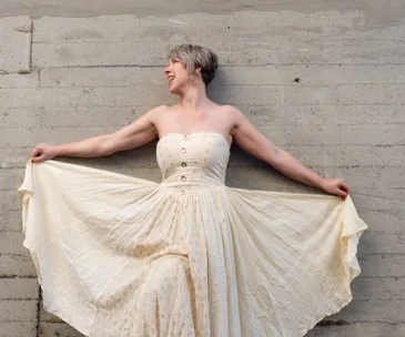 Woman in a cream dress, arms stretched wide, smiling against a concrete wall.