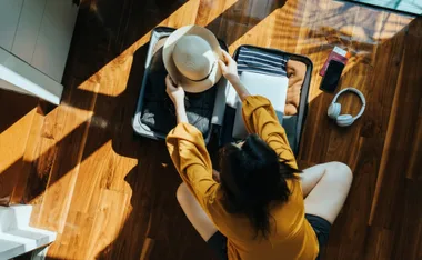 Person in yellow shirt packing a suitcase on a wooden floor, with a hat, passport, phone, and headphones nearby.
