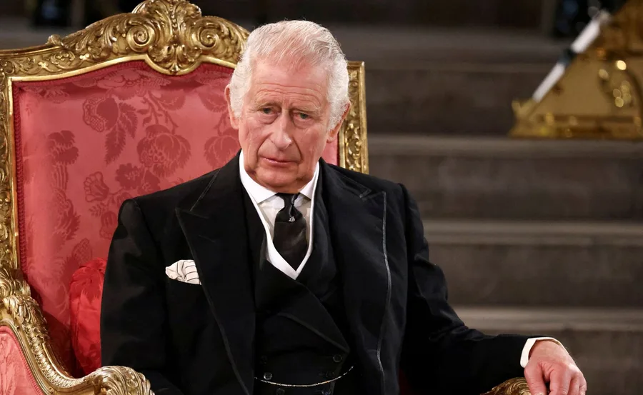 King Charles III sitting on an ornate red and gold chair, wearing a black suit and tie.