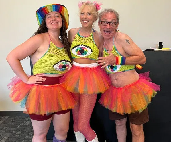 Three people in colorful outfits with crochet eyeball tops and rainbow tutus, standing together and posing indoors.