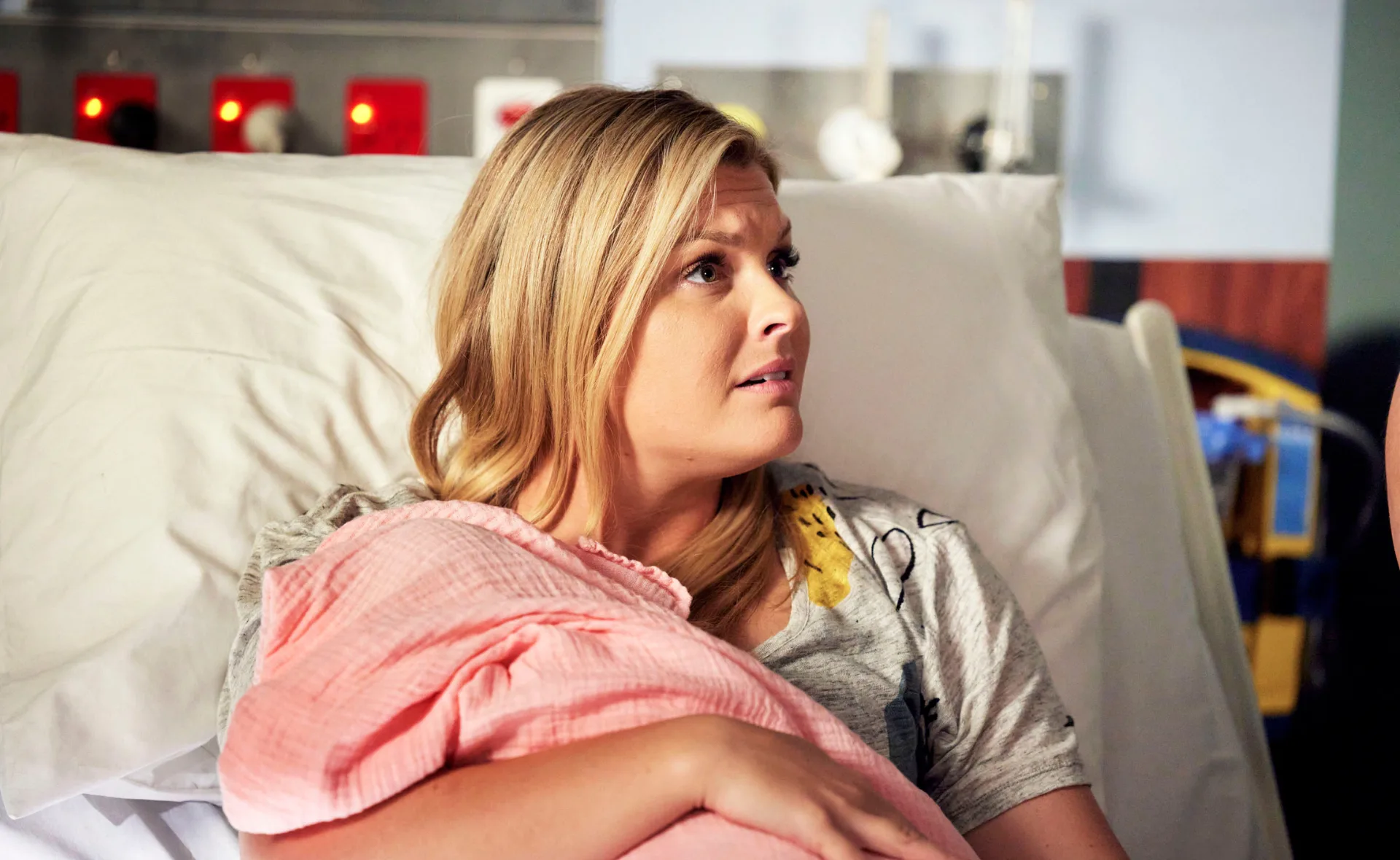 Woman with blonde hair sits in a hospital bed holding a baby wrapped in a pink blanket, looking pensive.
