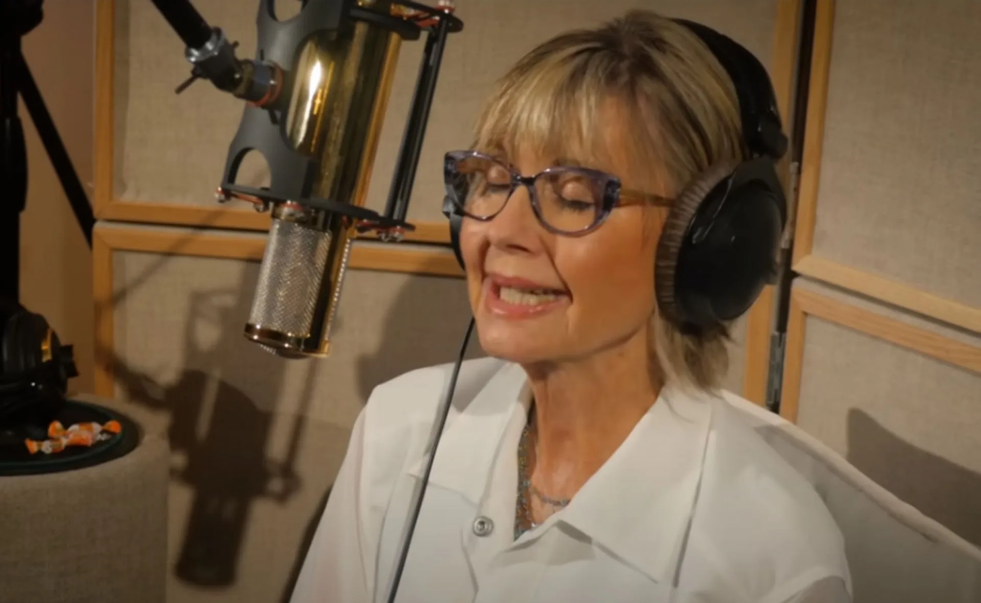 A woman in glasses and headphones singing into a studio microphone.