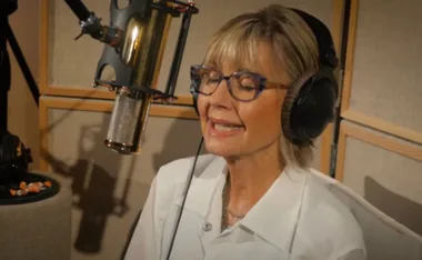 A woman in glasses and headphones singing into a studio microphone.