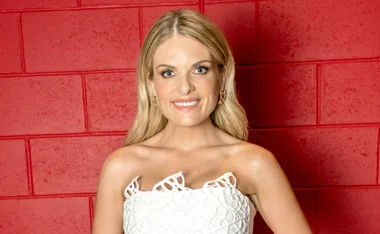 A woman with long blonde hair in a white dress smiles in front of a red brick wall.