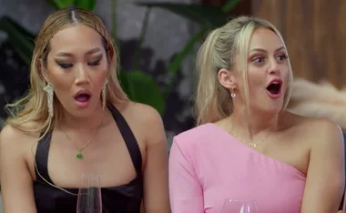 Two women sitting side by side, both with expressions of surprise. One wears black, and the other wears pink.