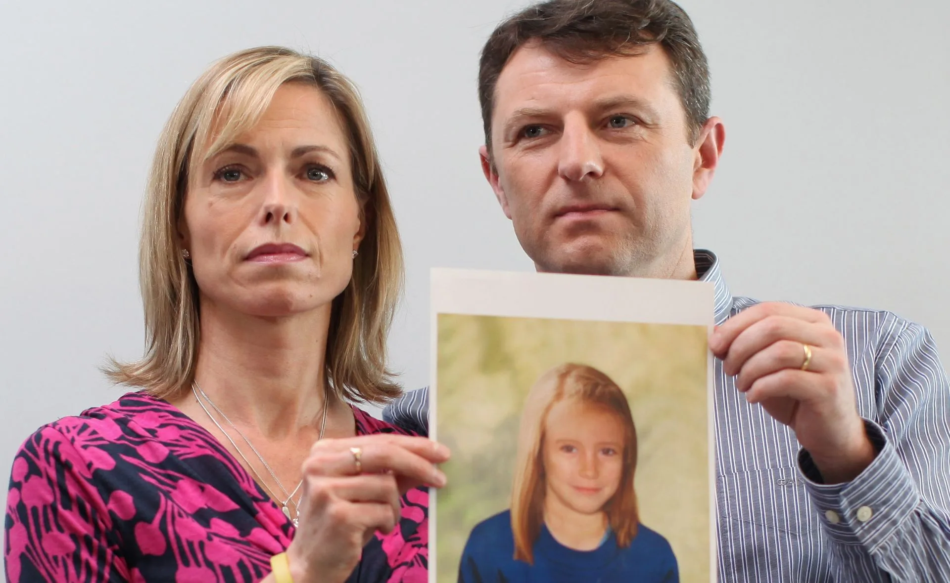 A man and woman hold a photo of a young girl with long blonde hair wearing a blue shirt.