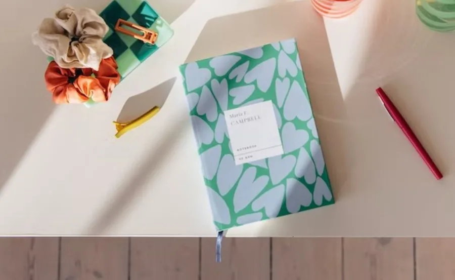 Notebook with heart design on desk, surrounded by scrunchies, a red pen, yellow hair clip, and checkered tray.