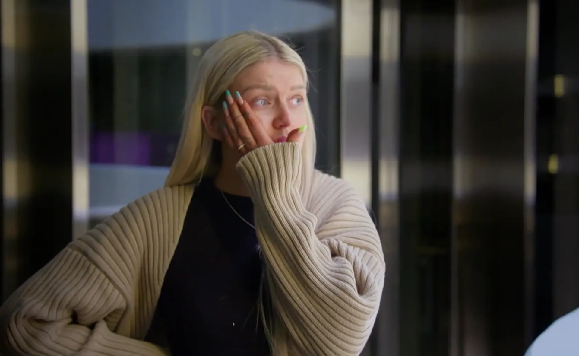 A woman with long blonde hair and a beige cardigan rests her hand on her face, looking off to the side.