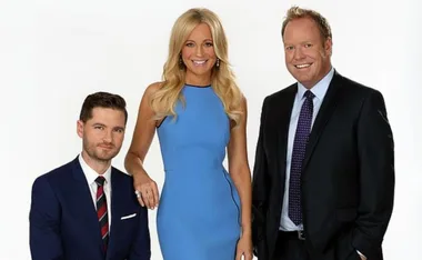 Three people in formal attire; one sitting, two standing, smiling against a plain background.