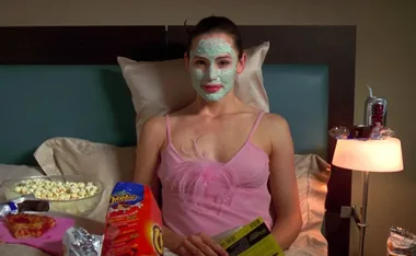 A woman in a pink top with a green face mask sits in bed with snacks like popcorn and chips around her.
