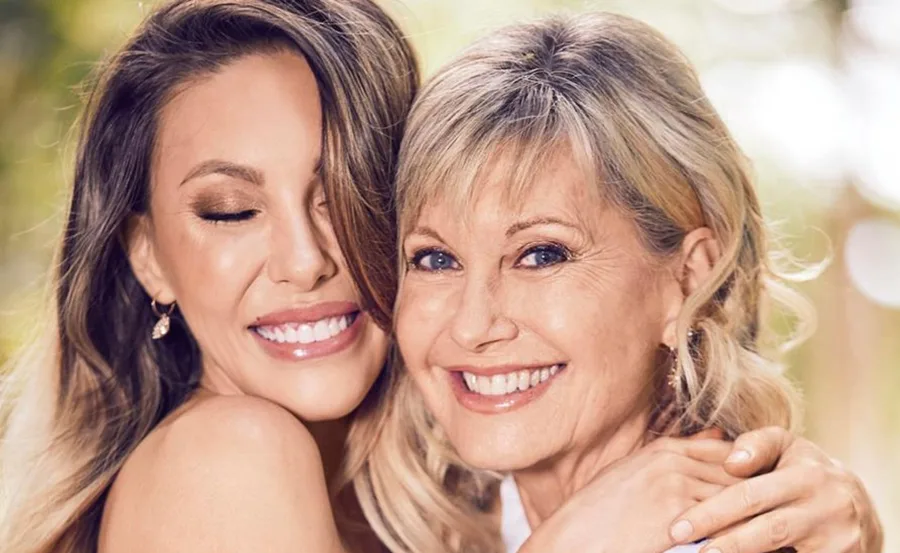 Two women smiling warmly, embracing each other, with a blurred outdoor background.